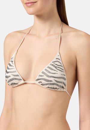 Vrouw draagt een beige bikinitop met zwarte zebraprintachtige stippen en dunne nek- en rugbandjes tegen een effen achtergrond.