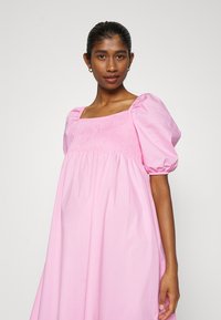 Vestido de algodão rosa com decote quadrado, mangas bufantes e detalhes pregueados no corpete, apresentando um corte solto e tecido fluido.