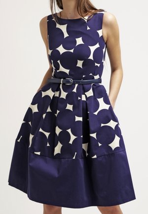Robe sans manches bleu marine et blanche à motifs, taille cintrée, jupe plissée et ceinture fine bleu marine, portée avec les mains dans les poches.