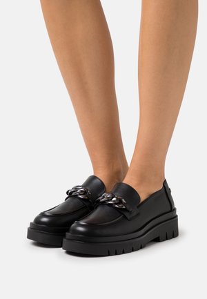 Mocassins en cuir noir avec une semelle épaisse et rainurée, agrémentée d'une chaîne décorative sur le dessus. Texture lisse avec un design à bout arrondi.