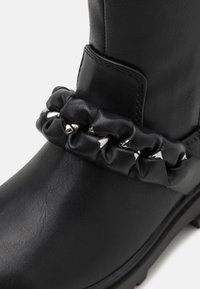 Bottine noire avec une texture en cuir lisse, bout rond, et une sangle nouée ornée de clous argentés autour de la cheville.