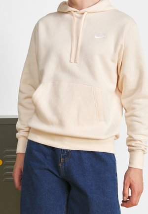 Person iført en lysebeige Nike hættetrøje med frontlomme og blå jeans, stående med den ene hånd afslappet ved siden af kroppen.