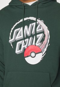 Sudadera verde oscura con el texto "Santa Cruz", un gráfico de Poké Ball en blanco y rojo, y cordones a juego en verde.