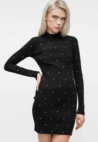 Egy alakra simuló, fekete bordás kötött ruhát viselő nő hosszú ujjakkal és apró ezüst szegecsekkel, az egyik kezét a csípőjén pihenve pózol.