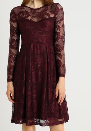 Kobieta nosząca bordową koronkową sukienkę do kolan z długim rękawem, wzorami kwiatowymi i dopasowaną talią.