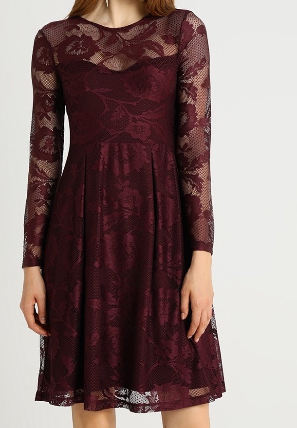 Femme portant une robe en dentelle bordeaux mi-longue, à manches longues, avec des motifs floraux et une taille cintrée.
