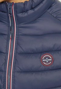 Tamnosinji puffer jakna s visokim ovratnikom, sa prednjim zatvaračem s crvenim i bijelim prugama te kružnim logom na prsima.