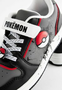 Schwarzer und grauer Sneaker mit einem weißen Riemen, der mit "POKÉMON" beschriftet ist, ausgestattet mit einem roten und schwarzen Pokéball-Abzeichen und strukturierten Akzenten. Weiße Schnürsenkel.