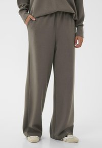 Person wearing loose-fitting taupe pants and matching sweatshirt, with hands in pockets and white sneakers visible below.