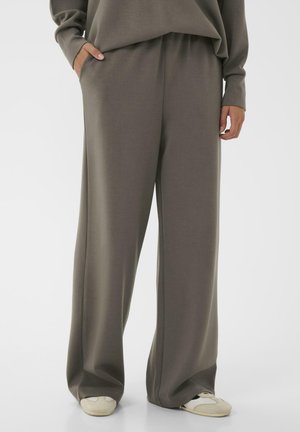 Personne portant un pantalon ample taupe et un sweat-shirt assorti, avec les mains dans les poches et des baskets blanches visibles en bas.