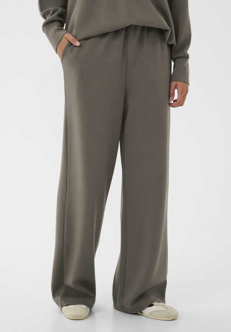 Person wearing loose-fitting taupe pants and matching sweatshirt, with hands in pockets and white sneakers visible below.