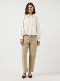 Vrouw in een witte geborduurde blouse met pofmouwen, beige rechte broek en zwarte pointy hakken, staand op een witte achtergrond.
