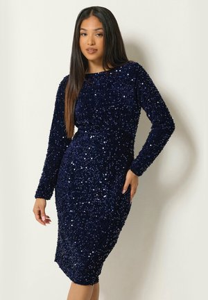 Femme portant une robe ajustée, manches longues, longueur genoux, bleu marine à sequins, debout devant un fond clair uni.