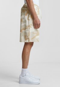Beige camo shorts made from cotton blend, featuring an elastic waistband and side pockets, paired with white sneakers and socks.