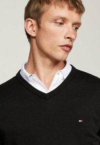 Schwarzer V-Ausschnitt-Pullover, der über einem weißen Hemd mit Kragen getragen wird; verfügt über ein kleines Markenlogo auf der Brust und eine glatte Textur.