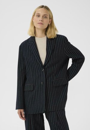 Blonde vrouw in een zwart gestreepte blazer en bijpassende broek met een beige top, staande met één hand op de heup tegen een effen achtergrond.