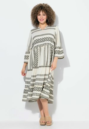 Lange, kaftan-stijl jurk in crème met zwarte geometrische patronen, met een tiered zoom en driekwart mouwen. Gedragen met beige plateau schoenen.