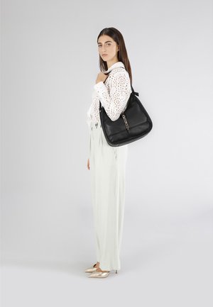 Borsa a tracolla in pelle nera con superficie texture, forma arrotondata e un accento decorativo in catena. Il modello indossa pantaloni larghi verde chiaro e una blusa bianca.