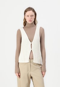 Weißes Rippentop mit V-Ausschnitt und einem Ausschnitt vorne, kombiniert mit einem braunen Langarmrollkragenpullover und beigefarbenen Hosen.