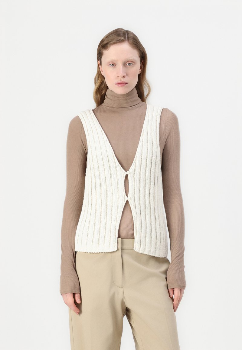 Weißes Rippentop mit V-Ausschnitt und einem Ausschnitt vorne, kombiniert mit einem braunen Langarmrollkragenpullover und beigefarbenen Hosen.