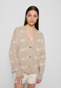 Beige cardigan met een V-hals en een oversized pasvorm, met een herhaalpatroon van "BIZU" in het wit en vier gouden knopen. Gewoven gebreid materiaal.