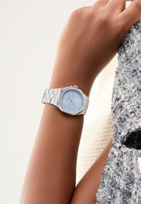 Srebrna osmostrana ura z svetlo modrim številčnico, srebrnimi kazalci in pasom iz nerjavečega jekla, nosena na zapestju ob teksturirani tkanini.