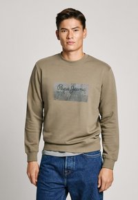 Sudadera verde oliva con cuello redondo y puños acanalados, presenta un logo gráfico impreso centrado en la parte delantera. Combinada con jeans azules.