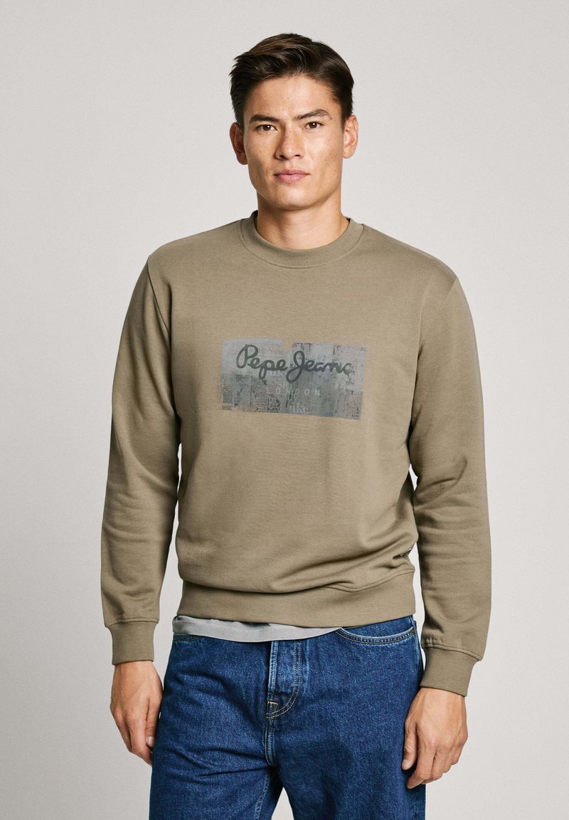 Sudadera verde oliva con cuello redondo y puños acanalados, presenta un logo gráfico impreso centrado en la parte delantera. Combinada con jeans azules.