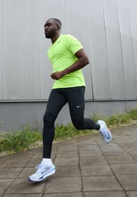Neon żółta koszulka sportowa, czarne legginsy z logo Nike, białe i niebieskie buty do biegania, plener z trawnikiem w tle.