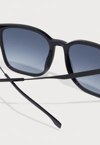 Ochelari de soare negri cu lentile gri gradient, având un design pătrat îndrăzneț și rame negre elegante. Temprile subțiri se extind de fiecare parte.