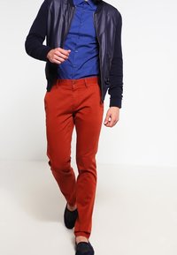 Námořnická kožená bomber bunda s žebrovanými rukávy, modrá košile s puntíky a těsně padnoucí kalhoty v barvě rzi. Černé slip-on boty dotvářejí outfit.