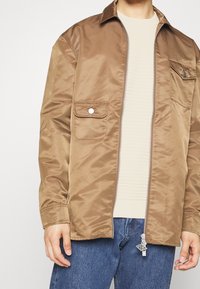Homme portant une veste en satin marron clair ouverte avec des poches avant, par-dessus un pull côtelé crème et un jean bleu, main détendue le long du corps.