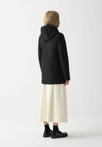 Juoda gobtuvinė striukė, pagaminta iš minkštos medžiagos, su tiesiu kirpimu ir minimaliu siūlių detalizavimu, derinama su balta sijonu ir juodais batais.