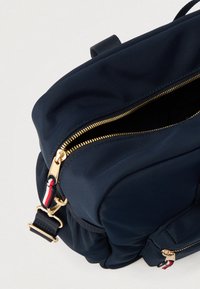 Σκούρο μπλε υφασμάτινο σακίδιο με χρυσά φερμουάρ, διακοσμημένο με λουρί από υφαντό ύφασμα με κόκκινες και λευκές λεπτομέρειες και κορυφαία λαβή για εύκολη μεταφορά.