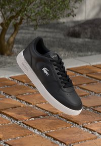 Sneakers negre din piele cu talpă albă. Prezintă un design cu șireturi, perforații pe vârful încălțămintei și logo-ul Lacoste în alb pe lateral.