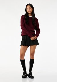 Suéter burdeos de punto trenzado con puños acanalados, combinado con una falda negra plisada. La modelo lleva calcetas negras hasta la rodilla y zapatos de plataforma.
