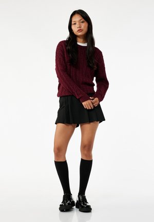 Burgundowa sweter z dzianiny warkoczowej z prążkowanymi mankietami, zestawiony z czarną spódnicą plisowaną. Modelka nosi czarne skarpetki do kolan oraz buty na platformie.
