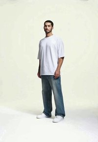 Homem de pé com expressão neutra, vestindo uma t-shirt branca larga, jeans azuis e ténis brancos contra um fundo claro.