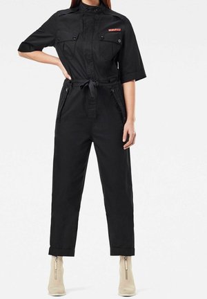 Combinaison noire à manches courtes, poches poitrine boutonnées, col mandarin et ceinture à nouer. Confectionnée dans un tissu lisse et léger.