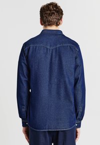 Camicia in denim blu con design della spalla posteriore, maniche lunghe con polsini abbottonati e orlo dritto. Tessuto liscio e cuciture uniformi.