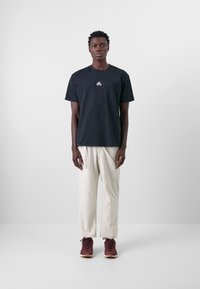Schwarzes Baumwoll-T-Shirt mit Rundhalsausschnitt und kleinem weißen Grafiklogo, kombiniert mit hellcremefarbenen, weiten Hosen und kastanienbraunen Sneakern.