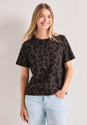 Młoda kobieta uśmiechnięta, ubrana w czarny i brązowy t-shirt w panterkę z kieszonką na piersi oraz jasnoniebieskie dżinsy, stojąca na tle jednolitej ściany.