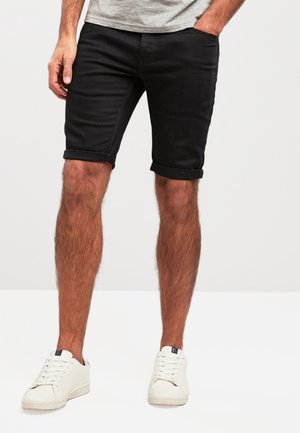 Homme portant un short noir retroussé, des baskets blanches et une chemise grise avec une main dans la poche, sur un fond uni.