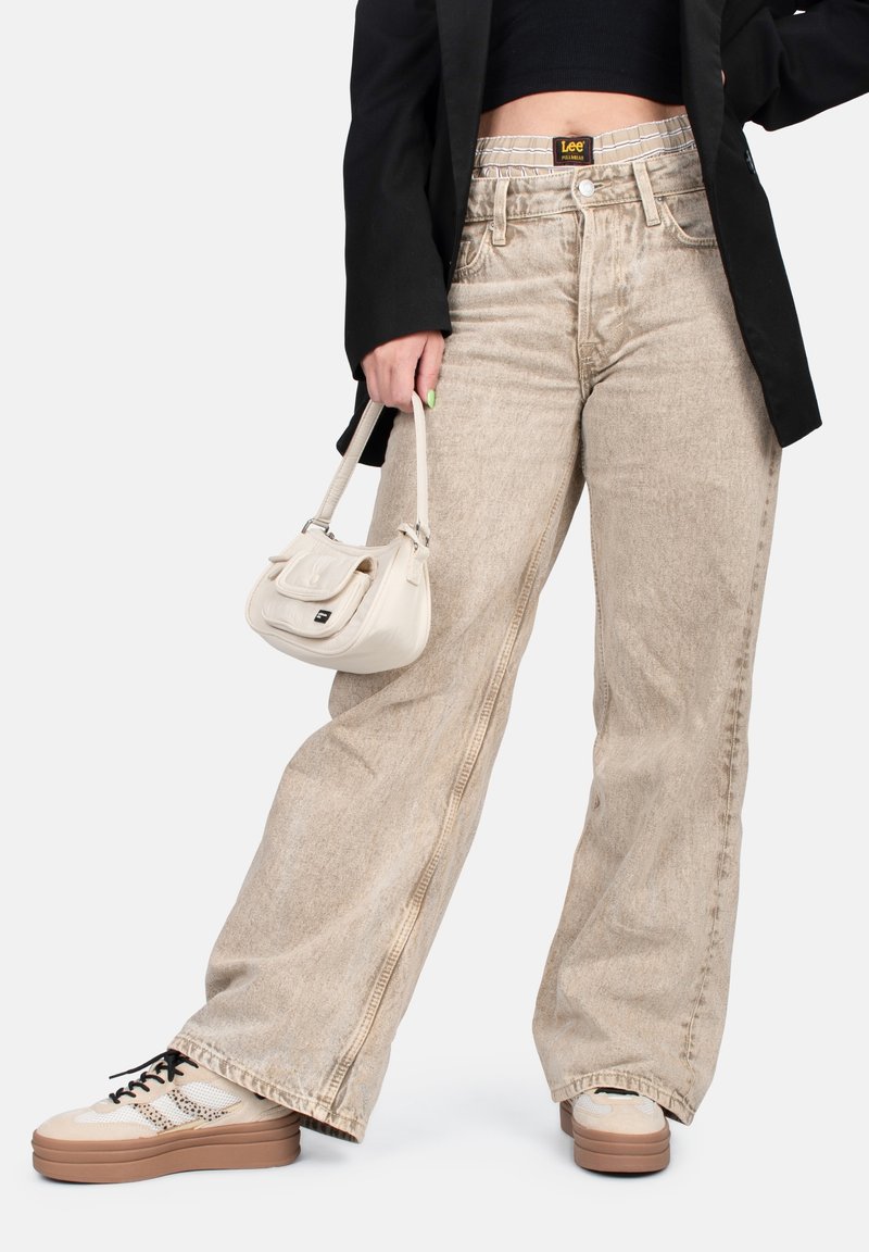 Personne portant un jean beige ample avec une étiquette de taille visible, un haut court noir, un blazer noir, tenant un petit sac à main beige et des baskets beiges.