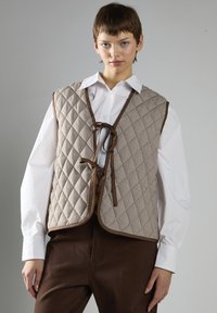 Quilted brun og tan vest med et diamantmønster, der har en snørelukning og kontrasterende kant. Båret over en hvid skjorte med knapper.