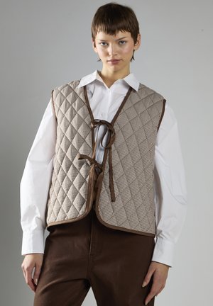 Quilted brun og tan vest med et diamantmønster, der har en snørelukning og kontrasterende kant. Båret over en hvid skjorte med knapper.
