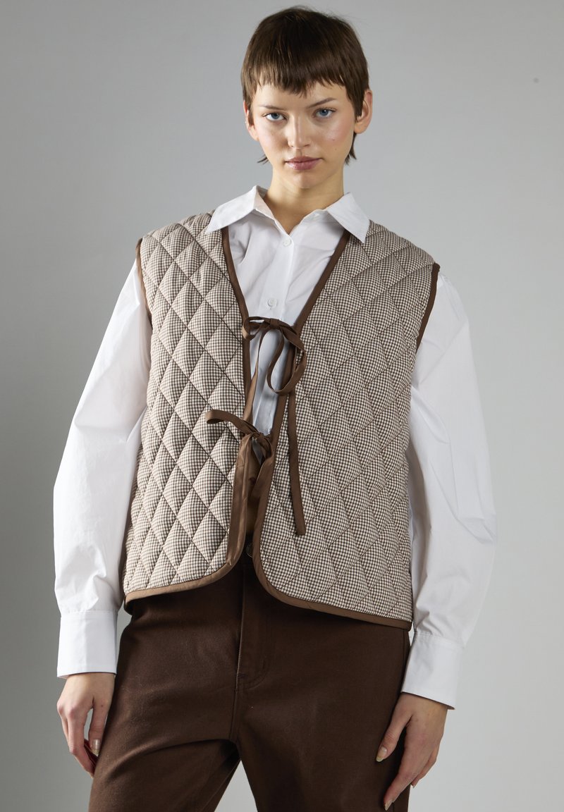 Quilted brun og tan vest med et diamantmønster, der har en snørelukning og kontrasterende kant. Båret over en hvid skjorte med knapper.