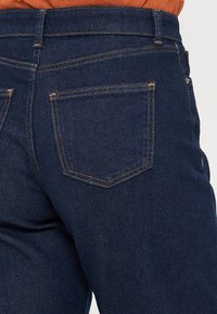 Tmavě modré džíny z denimu s hladkou texturou, s klasickými zadními kapsami s viditelným švem a s páskem střední výšky.