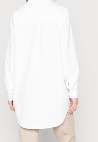 Chemise blanche à manches longues avec boutons sur le devant, coupe décontractée, empiècement au dos et texture subtile. Présente un ourlet arrondi et des poignets boutonnés.