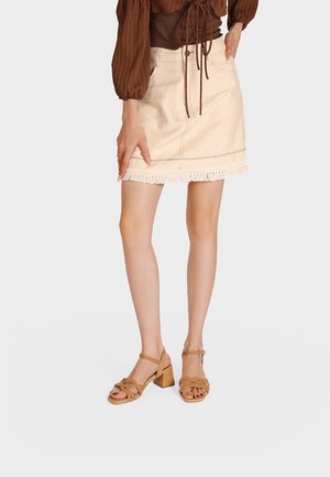 Donna che indossa una minigonna beige con orlo a frange, maglia marrone a maniche lunghe e sandali con tacco largo, punta aperta e intrecciati color cuoio, in piedi davanti a uno sfondo bianco.